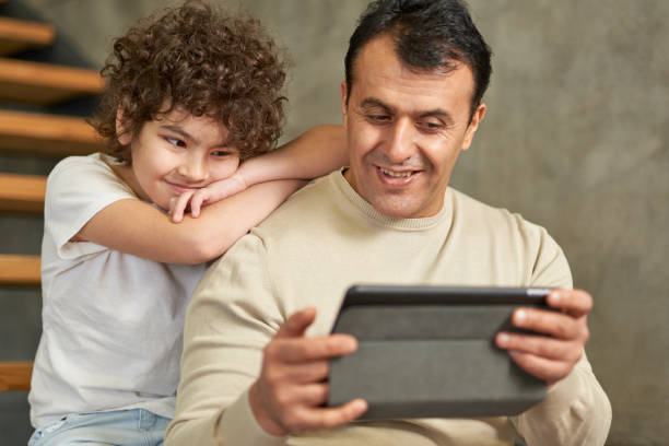 istock Middle aged latin father looking cheerful, holding tablet pc while spending time together with his son, sitting on the stairs at home 1311697383
