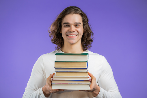 Student holds stack of university books from library on violet background in studio. Man smiles, he is happy to graduate. High quality photo