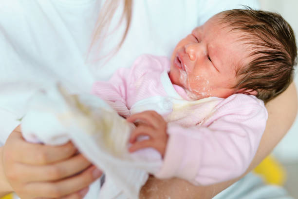 caucasian woman mother hold newborn infant girl vomiting milk after eating - baby overfeeding puke milk in hands of her mother at home - new life nursing and growing up concept stock photo