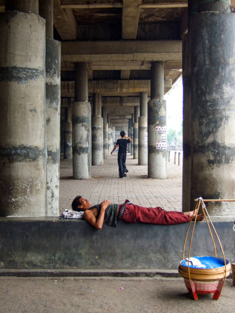 Homeless in Jakarta Vertical photo of two men, one sleeping on a cement ledge, the other walking away into the distance, under a large cement overhead bridge in a slum area of North Jakarta near the Old Port. A cane basket containing street food for sale is on the ground next to the sleeping man. jakarta slums stock pictures, royalty-free photos & images