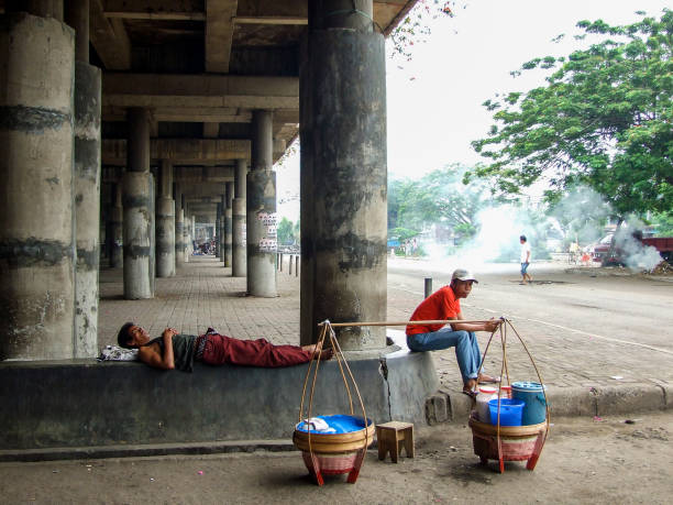 Poverty - Jakarta Horizontal photo of two men, one sitting, one sleeping on a cement ledge, under a large cement bridge in the city near the Old Port, Jakarta. Two cane baskets filled with street food, with a carrying pole, are on the ground next to the men. jakarta slums stock pictures, royalty-free photos & images