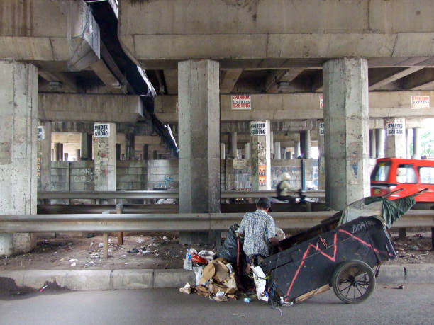 Horizontal photo of a man collecting rubbish from the street and putting it into a small hand pulled two-wheeled cart under a large overhead bridge with cement pylons in North Jakarta near the Old Port.