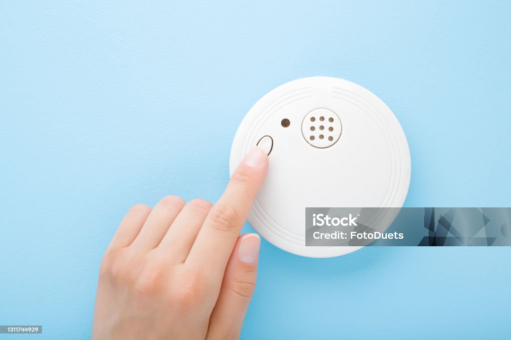 Young adult woman finger pushing button of new white plastic smoke alarm. Light blue table background. Pastel color. Closeup. Safety concept. Smoke Detector Stock Photo