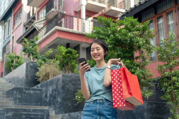 A tourist girl with a shopping bag using cellular mobile smartphones on a sidewalk in a garden park with the big-city background. 5G wireless communication technology, Global network telecommunication