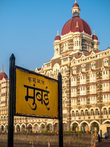 Mumbai, India - March 7, 2021 : Mumbai Signboard in the Marathi Language with Heritage Grand-class five-star hotel Taj, next to the Gateway of India.