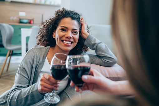 Shot of two happy women sitting on the floor talking and drinking wine. Smiling friends toasting with red wine at home. Two female friends relaxing at home with glass of wine.