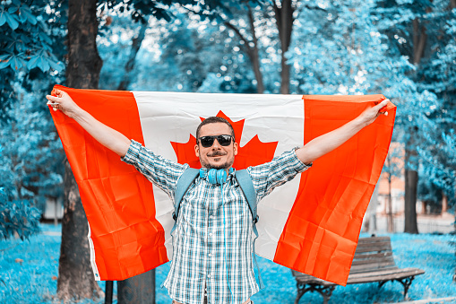 Young Handsome Tourist is Enjoying in Summer Day While Walking Through the Park With Colorful Trees, and Enjoying in Music Using Headphones While Holding a Flag of Canada.