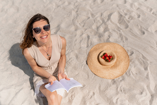 Happy smiling young woman sitting on the sand reading a book and looking up laughing. A straw hat with strawberry next to her. Shot from above.