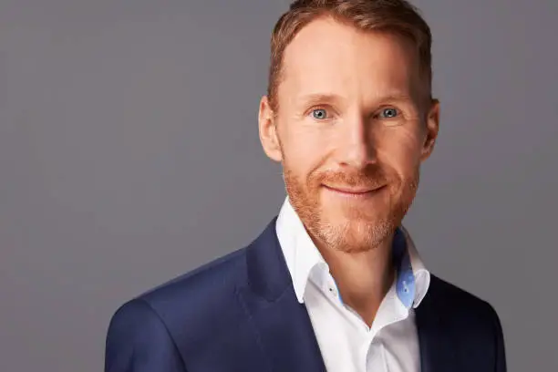 Portrait photos of a man in his forties with naturally red hair and a beard, dressed in a white shirt and blue jacket, photographed in the studio against a gray background. High resolution with copy space