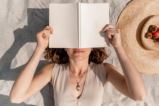 Attractive woman lying on the sand covering her face with a book. Shot from above.