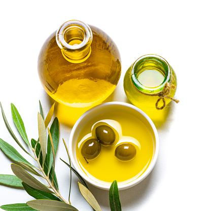 High angle view of two glass bottles and a ceramic bowl filled with extra virgin olive oil shot on white background. Green olives are in the bowl and an olive branch comes from the bottom of the frame. Predominant colors are yellow and white. High resolution studio digital capture taken with Sony A7rII and Sony FE 90mm f2.8 macro G OSS lens