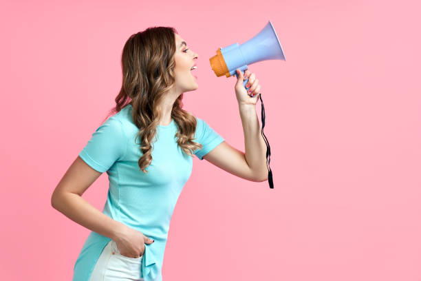 Pretty young woman shouting in megaphone on pink background stock photo