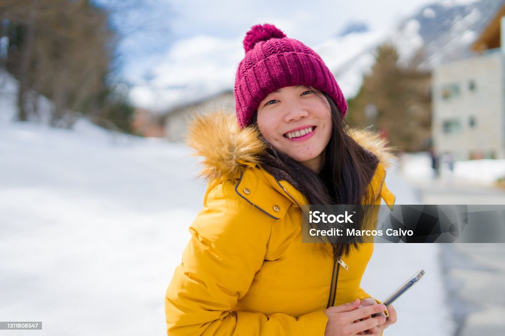 winter holidays in Swiss Alps - young beautiful and happy Asian Chinese woman holding mobile phone on snow landscape smiling cheerful in the cold mountain"n Ski Stock Photo