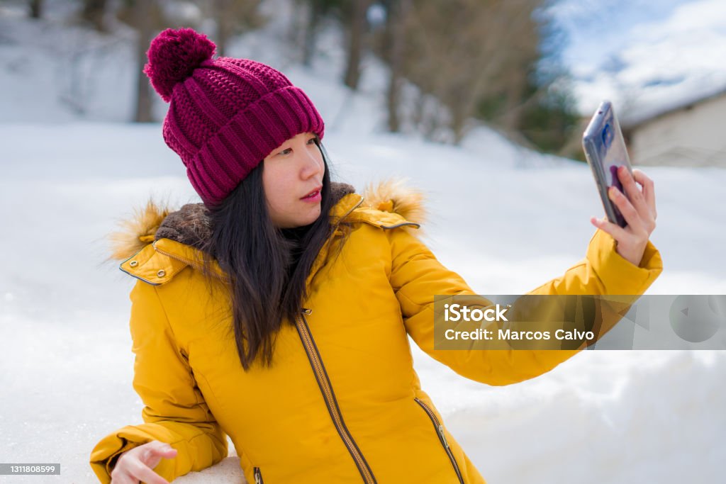 winter holidays in Swiss Alps - young beautiful and happy Asian Chinese woman holding mobile phone on snow landscape smiling cheerful in the cold mountain"n Japanese Ethnicity Stock Photo