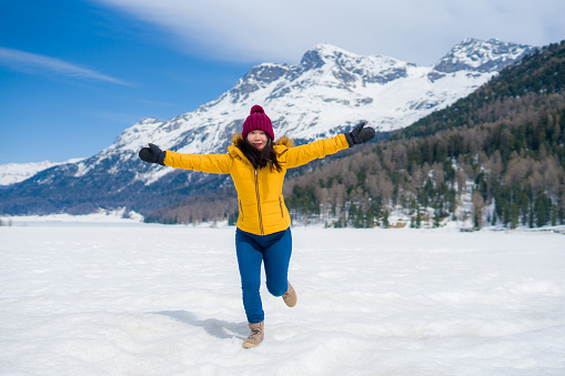 young happy and cheerful Asian Japanese woman in winter jacket and hat running playful on frozen lake covered in snow at Swiss Alps mountains enjoying holiday travel
