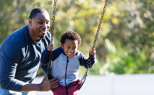 A father and son having fun together on the playground. The little boy, 3 years old, is sitting on a swing and dad is standing behind him, pushing. The child is mixed race Hispanic, African-American and Native American.