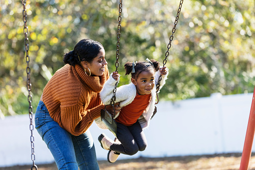 A mother and daughter having fun together on the playground. The little girl, 3 years old, is sitting on a swing and mom is standing behind her, pushing. They are mixed race Hispanic, African-American and Native American.