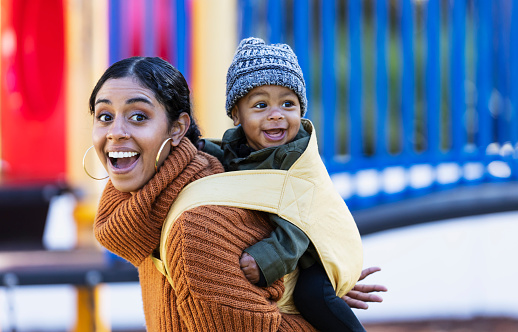 A mid adult woman in her 30s with her 7 month old baby boy on a playground. He is on her back in a baby carrier and they are both looking to the side, smiling. They are mixed race Hispanic, African-American and Native American.