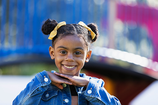 Headshot of a beautiful 5 year old mixed race girl outdoors on a sunny day at the playground. She is looking at the camera with a confident smile, hands under her chin. She is mixed race Hispanic, African-American and Native American.