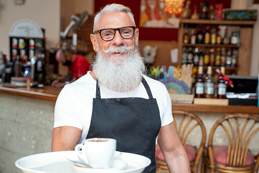 Bearded senior man in apron looking at camera during work in cozy coffee house, smiling.