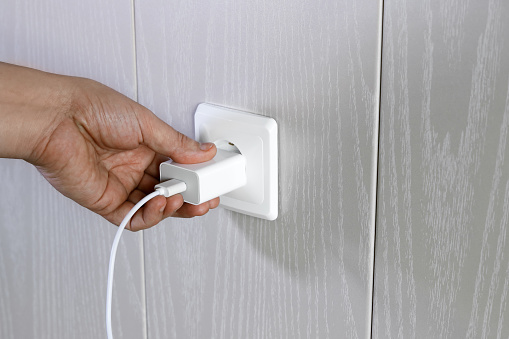 Hand turns on, turns off charger in electrical outlet on wall, close-up with copy space