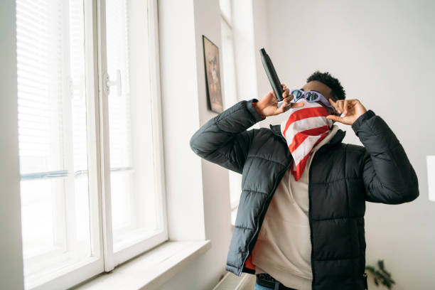 A man holding a microphone up and singing while wearing a stars and stripes face covering and sunglasses