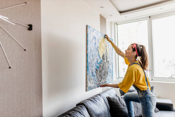 giovane donna appendere immagine d'arte a parete e decorare soggiorno - decorare foto e immagini stock