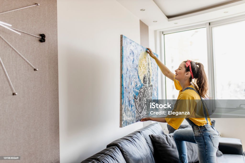 Young woman hanging art picture on wall and decorating living room Painting - Art Product Stock Photo