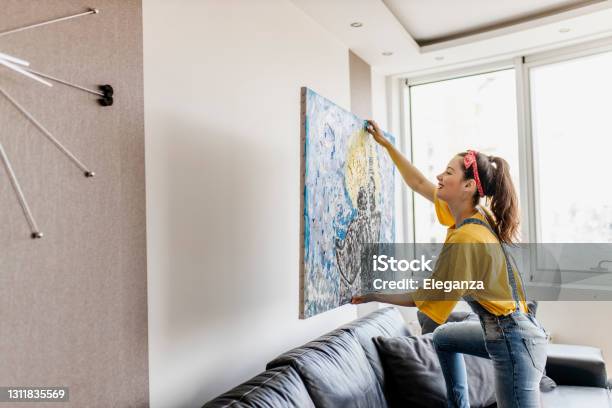 Young Woman Hanging Art Picture On Wall And Decorating Living Room Stock Photo - Download Image Now