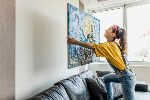 Young woman hanging art picture on wall and decorating living room
