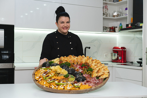 Charming and proud female chef showing of the appetizer plate, with a tasty food, she made from her modern kitchen