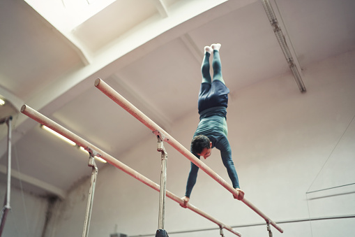 Focused and determinated young male athlete, a gymnast performing the routine on a parallel bar, during his training in the indoor gymnastic court