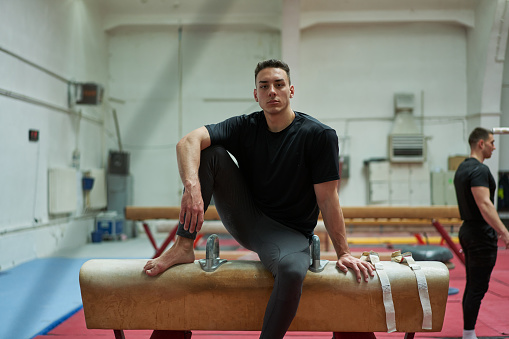 Portrait of a confident and serious young male gymnast sitting on a pommel horse, after he finished with a training