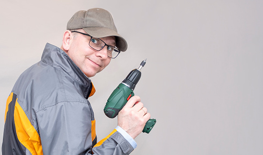 A Caucasian man with a cordless drill looks at the camera and smiles. Man construction profession on a gray background. Copy space.