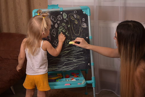Mom and daughter sit on the floor and draw with chalk on the blackboard
