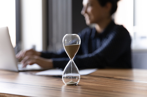 Close up hourglass measuring time, standing on wooden office table, Indian businesswoman working on background, efficiency, deadline and time management concept, busy employee using laptop