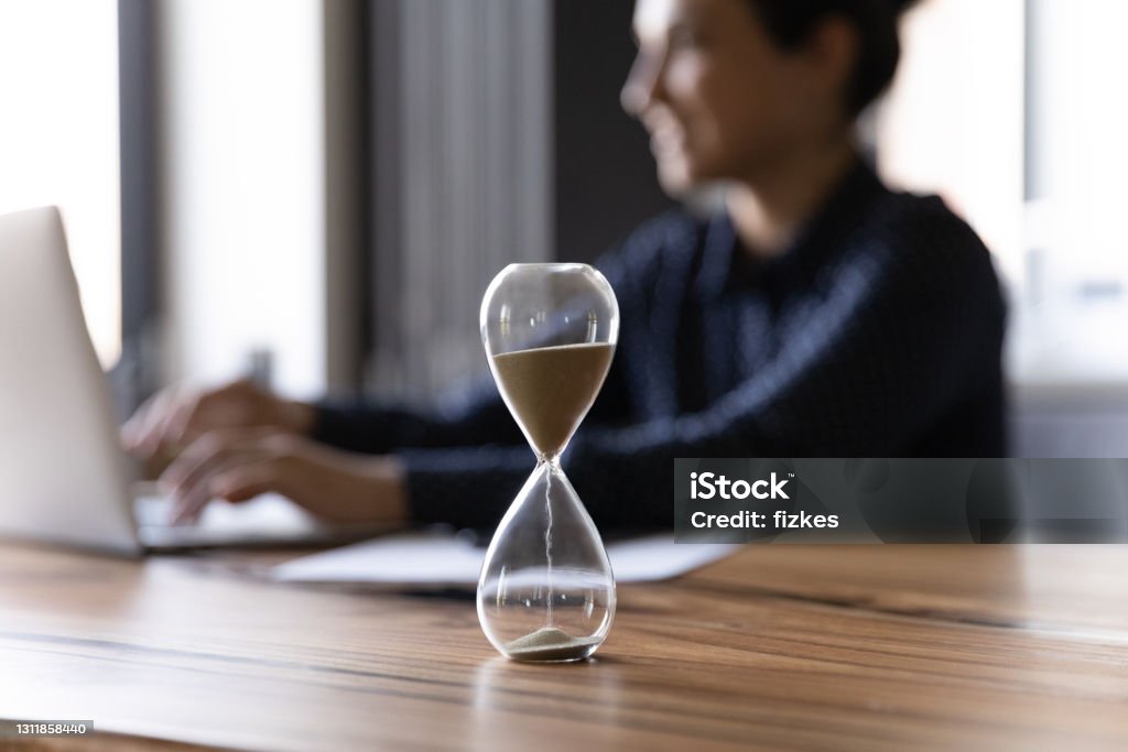 Close up hourglass measuring time, Indian businesswoman working Close up hourglass measuring time, standing on wooden office table, Indian businesswoman working on background, efficiency, deadline and time management concept, busy employee using laptop Time Stock Photo