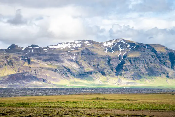 Landscape view near Gerduberg Basalt Columns in Snaefellsnes Iceland with green grass on summer day and clouds in sky and snowcapped volcanic mountains