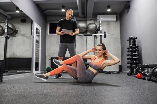 Measuring the success of sports training and motivation Woman in sportswear does sit-ups and abdominal exercises at the gym, while a trainer observes her training and enters data into a digital tablet