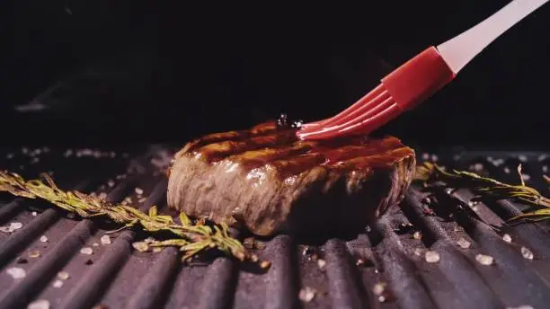 Photo of Delicious juicy meat steak cooking on grill. Prime rare roast grilling beef. Electric roaster, rosemary black pepper, salt. Silicone cooking brush.