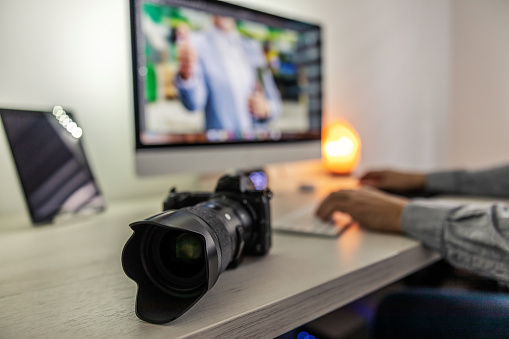 A photo zoom of the professional black camera on a neatly furnished desk. A man sits on a chair and type on the keyboard until he successfully completes the job. Professional photographer, freelance