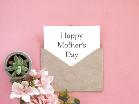 Happy Mothers Day letter in envelope and flowers around it on pink background.\nHappy Mothers Day letter.