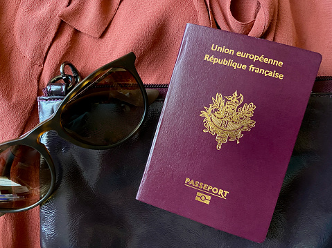 Horizontal high angle closeup photo of a French passport, a pair of sunglasses, a handbag and a partially seen item of women's clothing.