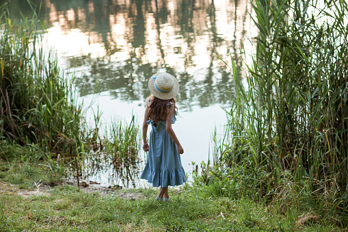 A cute girl in a long blue dress and straw hat stands on the shore of the lake. Back of a child in the public park with water, bushes, green and dried grass in summer. Young beautiful lady on walking.