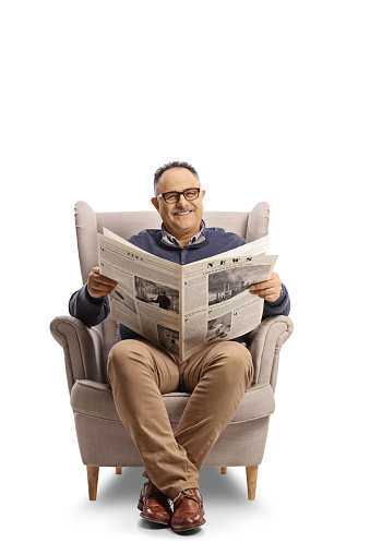 Cheerful mature man reading a newspaper and smiling seated in an armchair isolated on white background