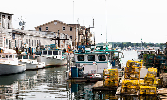 Portland, Maine, USA - 20 July 2019: Commercial fishing boats docked in a canal with colorful lobster traps stacked on the dock behind buildings.