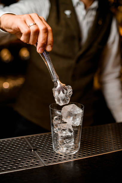 bartender holds piece of ice with tongs over glass on bar counter - transparent holding glass focus on foreground imagens e fotografias de stock
