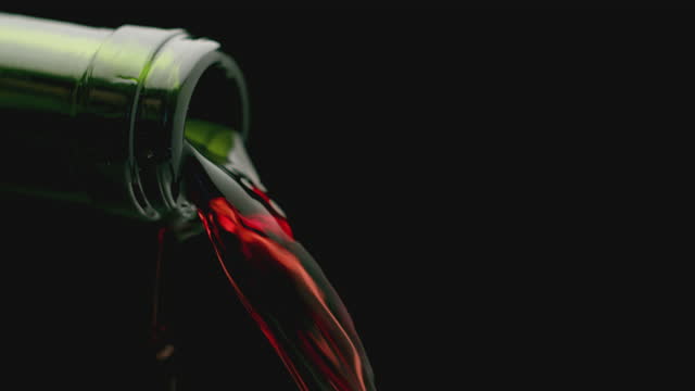 The slow tilt of the green bottle from which the wine is being poured. Slow action of pouring red wine out of a bottle, on a black background