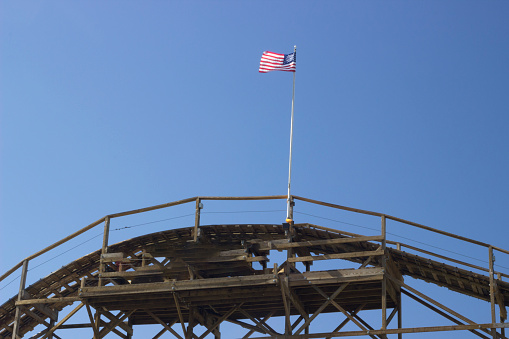 A United States flag flies high above the top of a wooden roller coaster. No ride cars are visible. The flag is fully unfurled to the left in a strong wind. Behind it is blue sky.