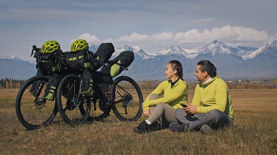 The man and woman travel on mixed terrain cycle touring with bikepacking. The two people journey with bicycle bags. Sport bikepacking, bike, sportswear in green black colors. Mountain snow capped.